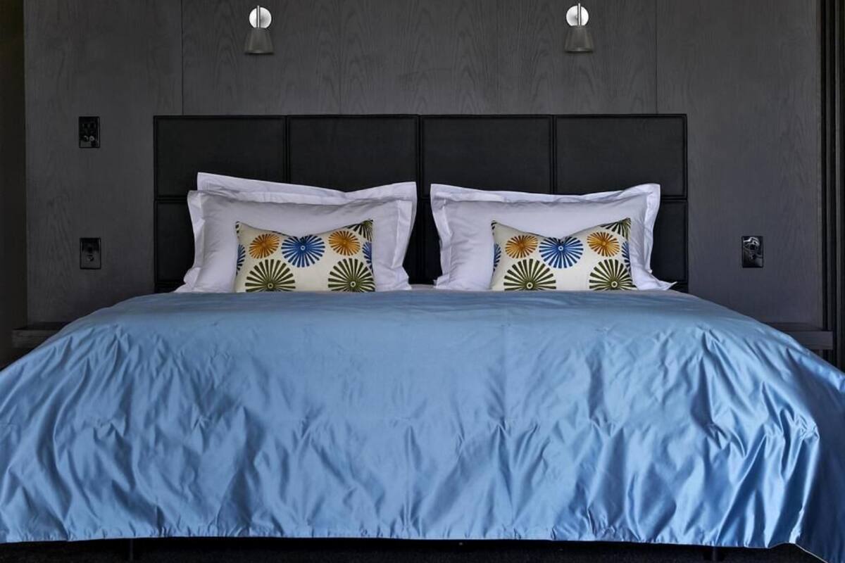 A double bed with a blue bedspread and decorative pillows, set against a dark wood panel wall with two wall-mounted lights.