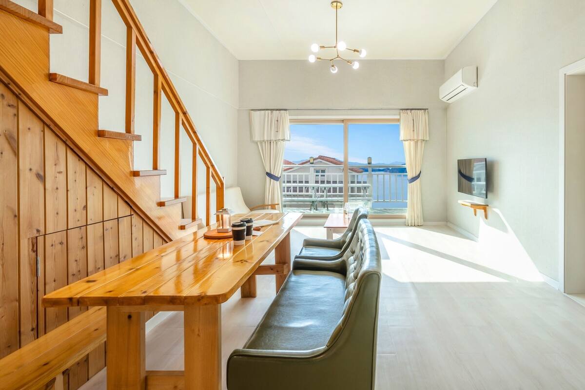 Ein modernes Hotelzimmer mit einer Holztreppe, einem Essbereich mit einer Bank, einem an der Wand befestigten Fernseher und Blick auf einen Balkon mit Geländer.