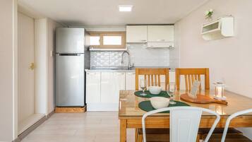 Une cuisine compacte équipée d’un réfrigérateur, d’un évier et de meubles de rangement en bois. On y trouve une table à manger avec des chaises et une petite lampe de table.