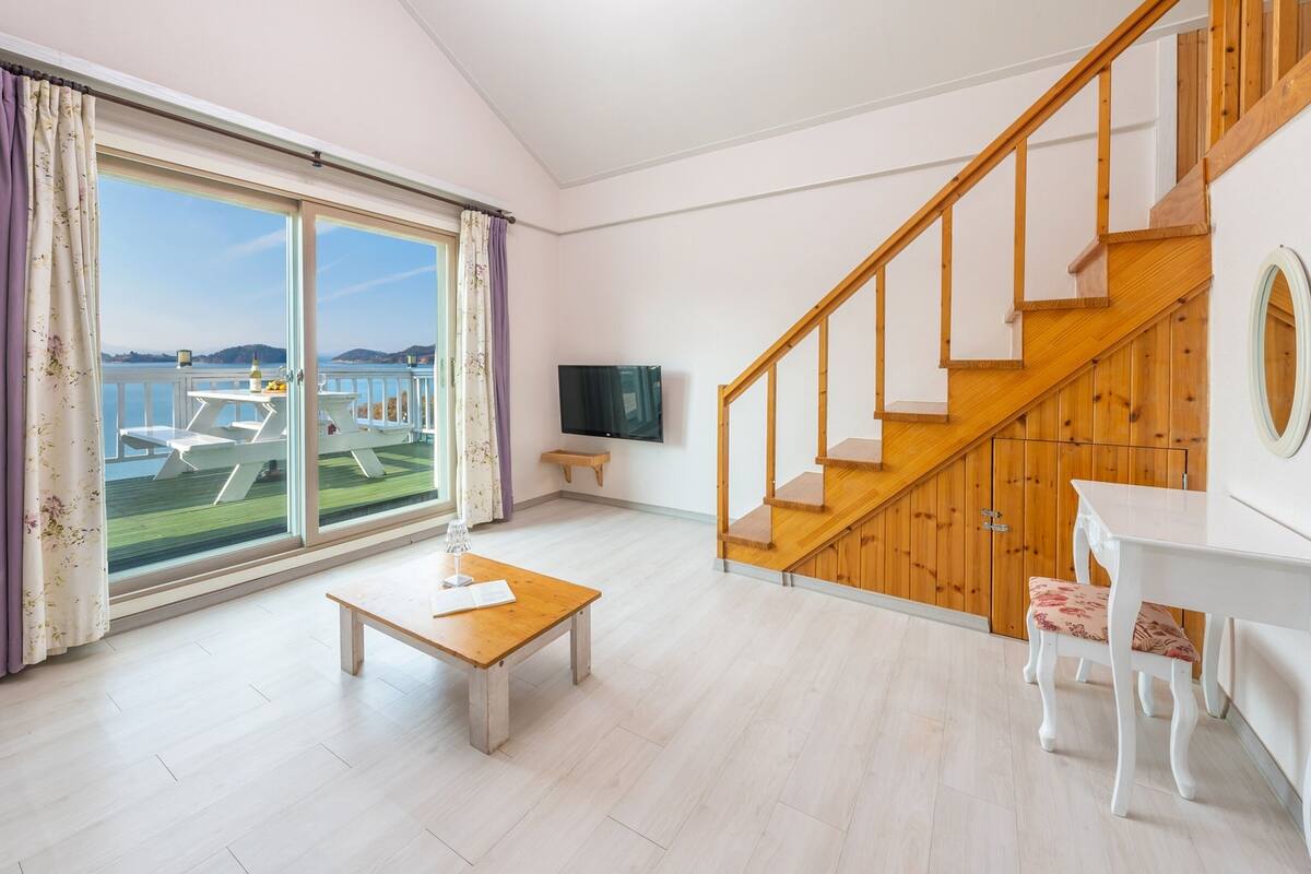 Ein geräumiges Wohnzimmer mit großen Schiebegläsern zum Balkon, einer Holztreppe, einem Flachbildfernseher und einem kleinen Tisch mit Stuhl.
