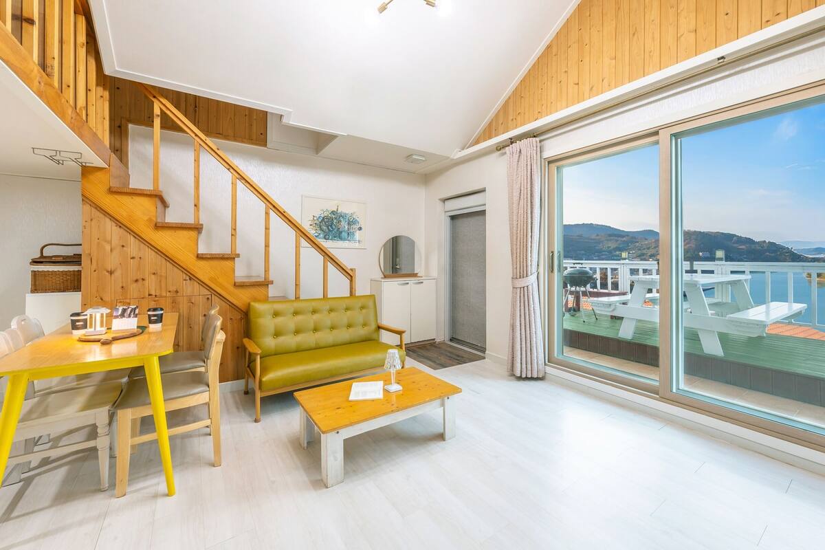 Ein modernes Wohnzimmer mit einer Holztreppe, einem grünen Sofa, einem Esstisch mit Stühlen und einer großen Schiebeglasdoor, die auf einen Balkon mit Blick auf Wasser und Hügel führt.