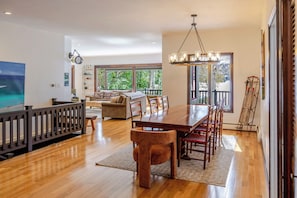A spacious dining area with a wooden table and chairs, a chandelier, and large windows offering a view of the outdoors.