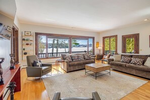 A spacious living room with a large window, a sofa set, a coffee table, and a wooden floor. - Dreamy Mirror Lake house retreat 5-Bedroom. 5 min walk to village (Lake Placid)