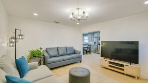 Un salon moderne avec un canapé gris, un téléviseur à écran plat posé sur un meuble en bois, un pouf rond et un lustre.