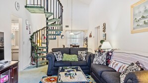 A living room with a dark blue sofa, a glass coffee table, and a colorful rug. A spiral staircase with a green railing leads to an upper level. - 273 Sea Woods Veranda 3BD - Keep Palm & Carry On (New Smyrna Beach)
