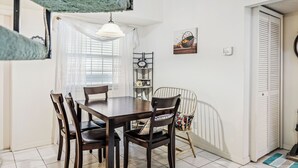 A dining area with a wooden table and chairs, a white ceiling light, a window with sheer curtains, a wall-mounted shelf with books and a clock, and a door with a white shutter. - 273 Sea Woods Veranda 3BD - Keep Palm & Carry On (New Smyrna Beach)