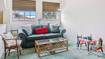 A cozy living room with a green sofa, a glass coffee table, and a small table with toys and a lamp.