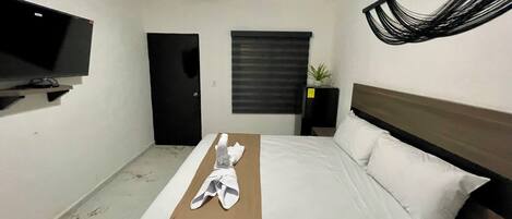 Una habitación de hotel con una cama, un televisor sobre un mueble, un ventilador de techo, una puerta, una ventana con persianas y una planta pequeña en una mesita de noche.