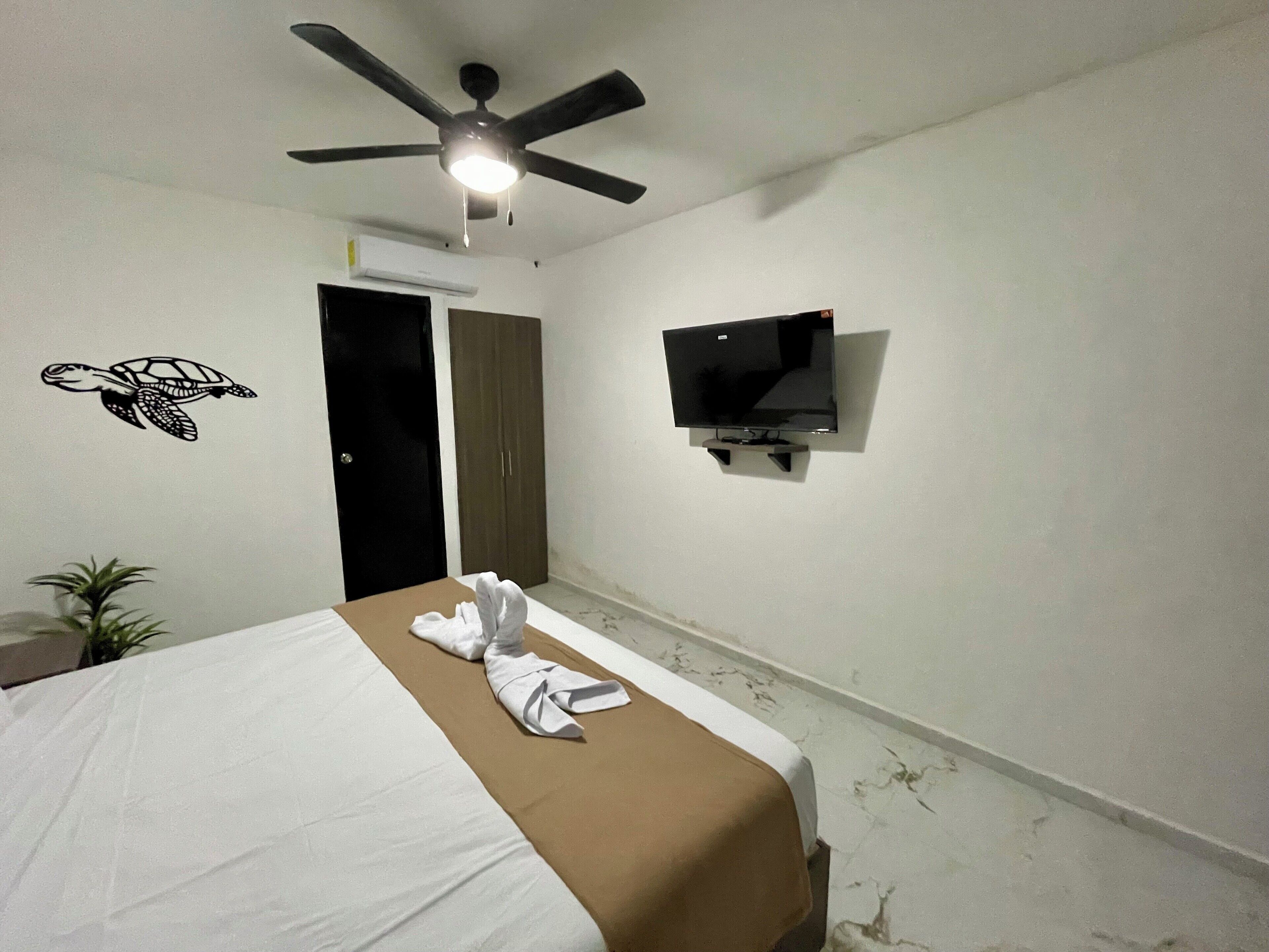A hotel room with a bed, a ceiling fan, a wall-mounted TV, and a towel on the bed.