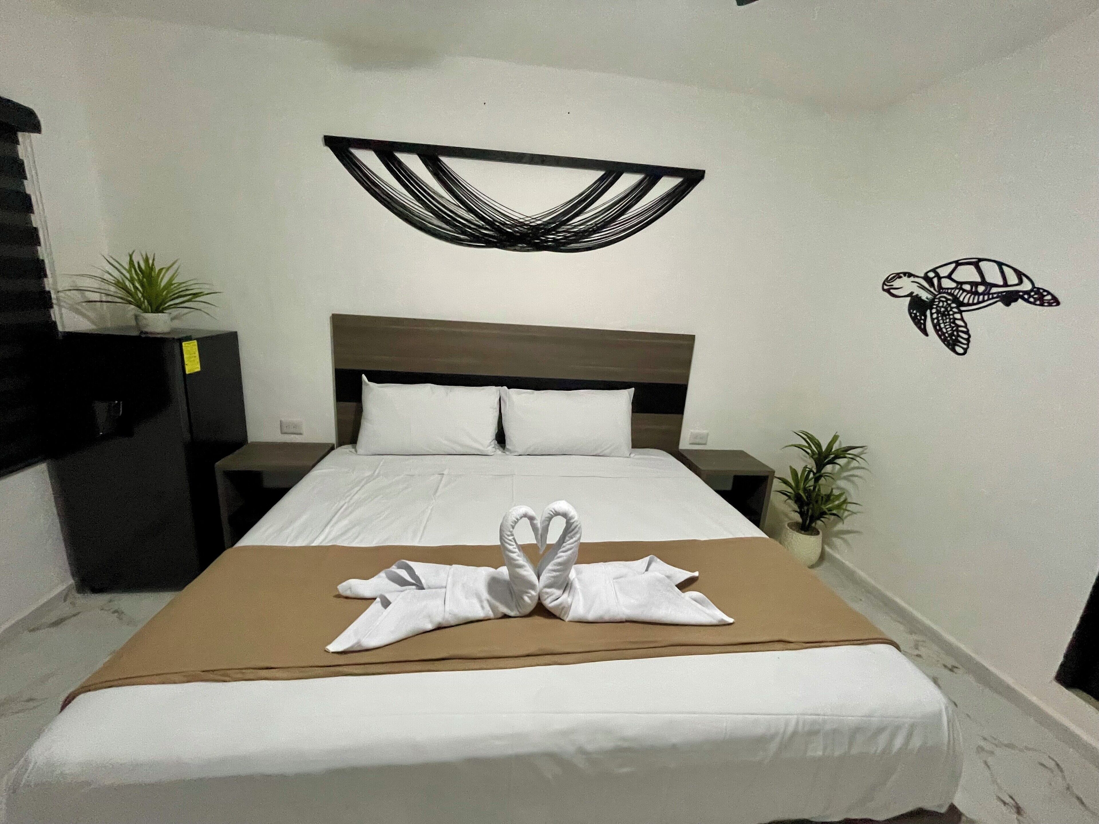 A neatly made bed with a brown bedspread and white pillows, a wall art of a turtle, and two potted plants on either side of the bed.