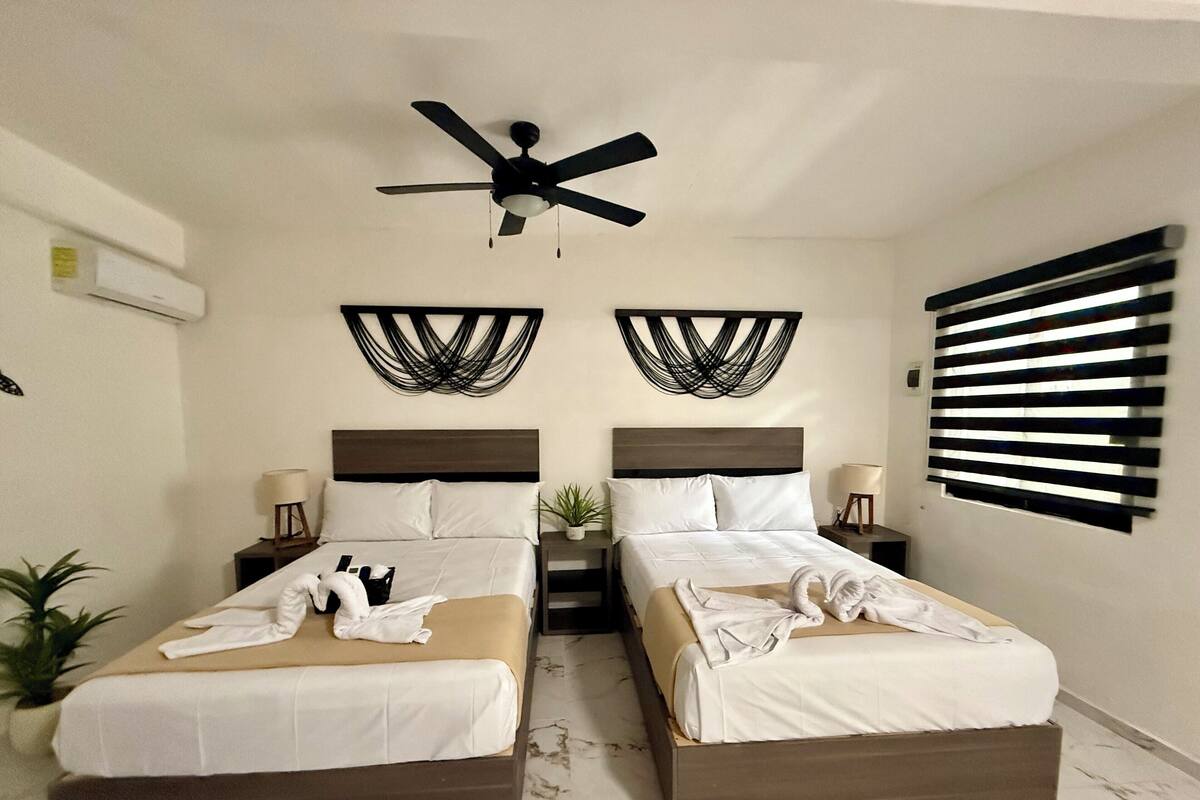 Egy szállodai szoba két ággyal, mennyezeti ventilátorral, a falra szerelt, absztrakt műalkotásokkal, valamint zsaluzatos ablakkal.