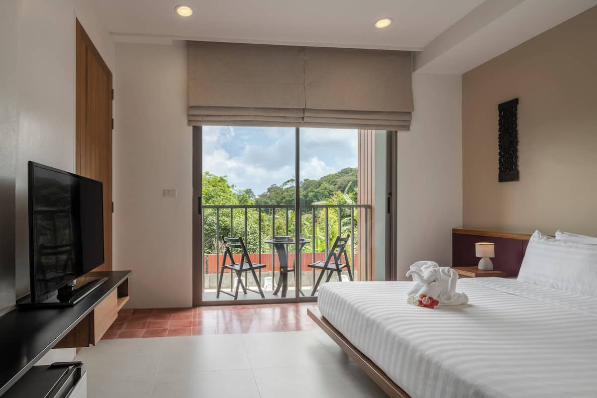 A hotel room with a large bed, a TV, a balcony with chairs, and a view of greenery.