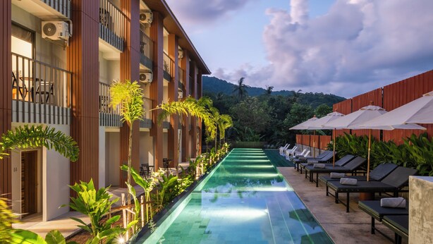 Un espace piscine doté de chaises longues et de parasols, entouré d’un bâtiment avec des balcons et des unités de climatisation.