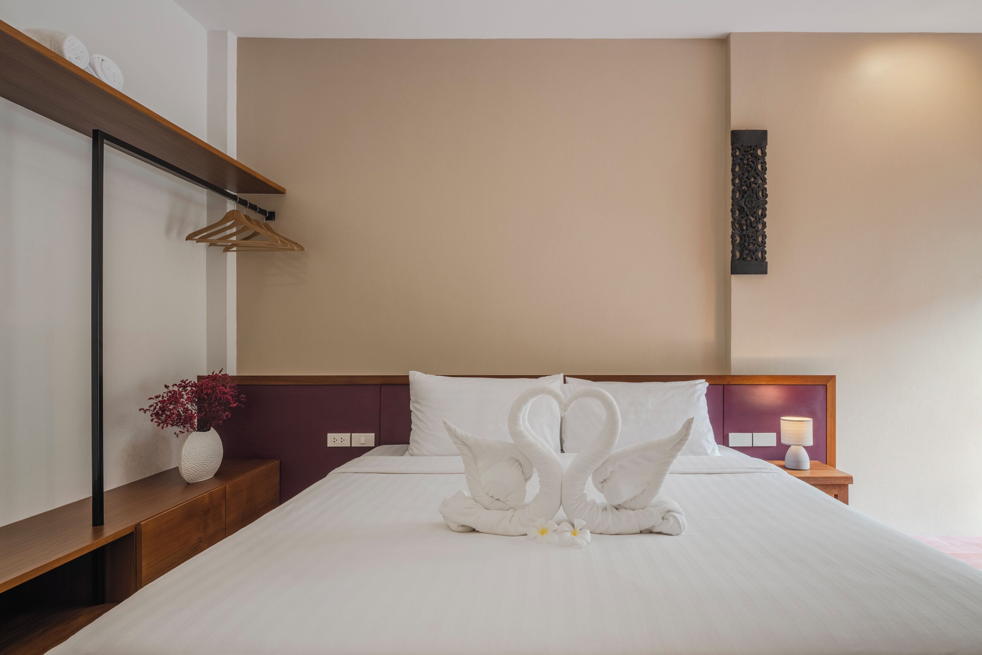 Ein Hotelzimmer mit einem ordentlich bezogenem Bett, einem Nachttisch aus Holz mit Lampe, einer Vase mit Blumen und einem an der Wand befestigten Regal mit Handtüchern.