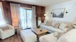 Un salon moderne comprenant un canapé, un fauteuil et un pouf, une table basse en bois et une grande fenêtre avec des rideaux.