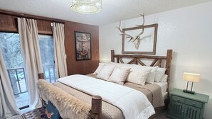 Une chambre avec un grand lit, une tête de lit en bois, un banc recouvert d’un plaid en fourrure, un tapis à motifs léopard, une table de chevet verte et un décor représentant un crâne de cerf.