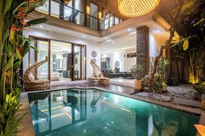 Area piscina interna moderna con piscina in vetro, mobili in vimini e una scalinata che conduce a un piano superiore.