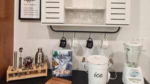 Una encimera de cocina con un menú de café, un libro sobre cócteles, un molinillo de café, una tabla de cortar, un cubo blanco con la etiqueta 'hielo' y una licuadora.