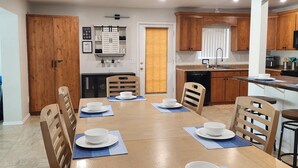 Un comedor con una mesa de madera dispuesta para cuatro personas, rodeada de sillas de madera. Se ve una cocina con armarios de madera y una ventana al fondo.