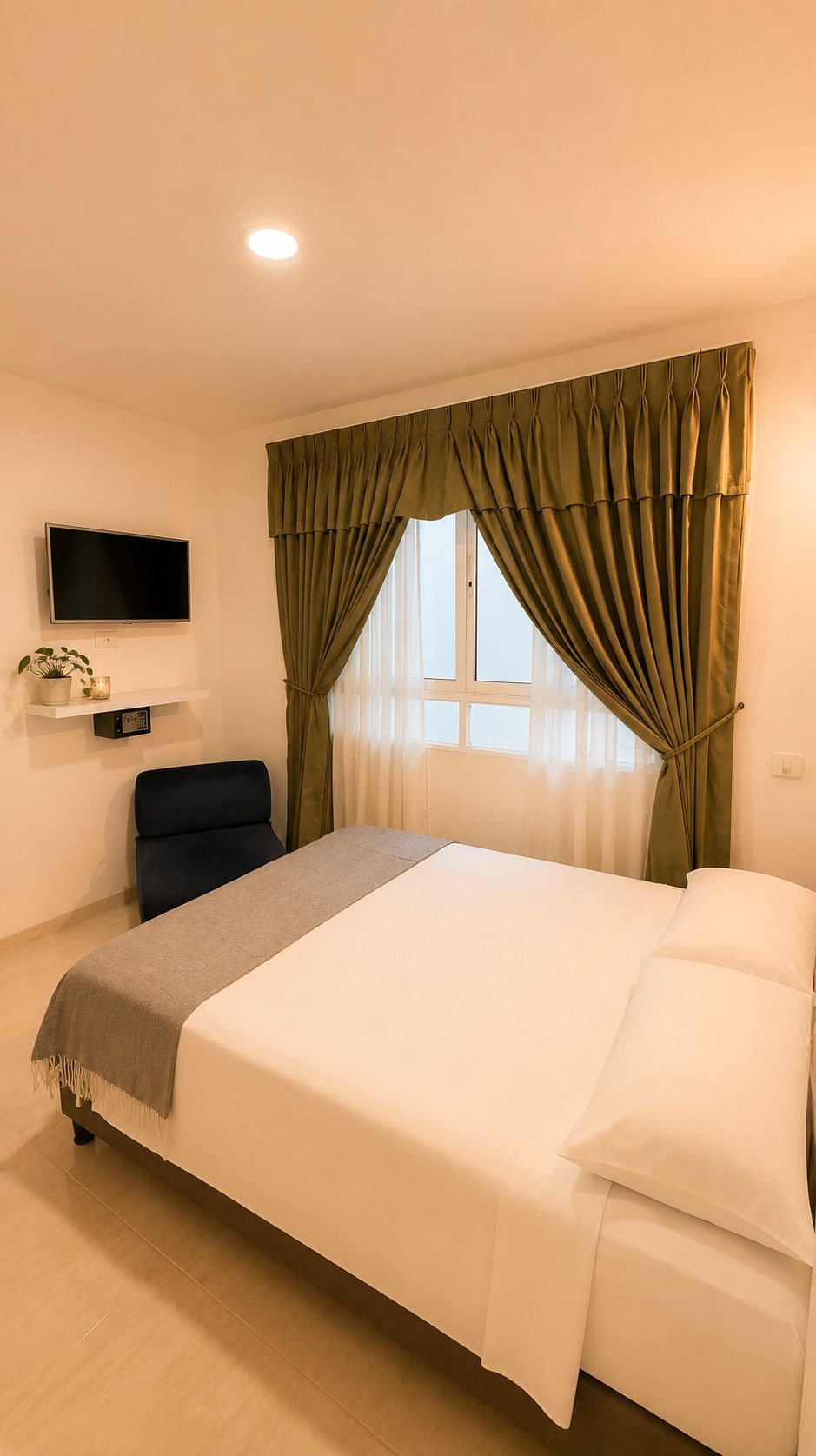 Habitación de hotel con cama, televisor montado en la pared, una silla y una ventana con cortinas.