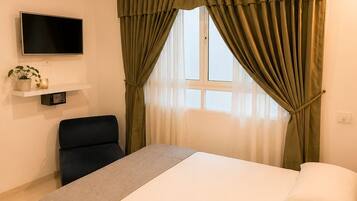 Habitación de hotel con cama, televisor montado en la pared, una silla y una ventana con cortinas.