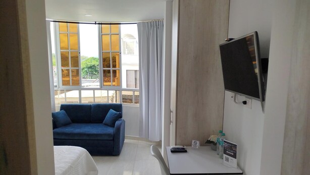 Habitación de hotel con cama, sofá azul, televisor, escritorio y control remoto, además de una ventana con cortinas.