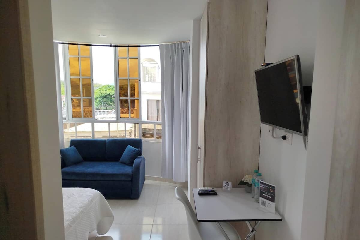 A hotel room with a bed, a blue sofa, a TV, a desk with a remote control, and a window with curtains.