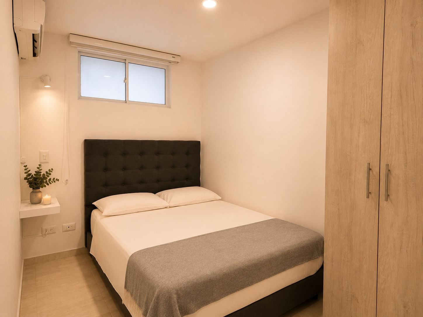 Ein Schlafzimmer mit einem Bett, einem Nachttisch mit Pflanze und Kerze, einem Fenster und einem Kleiderschrank.
