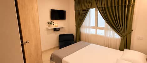 Un dormitorio con cama, una silla, un televisor en la pared y una ventana con cortinas.