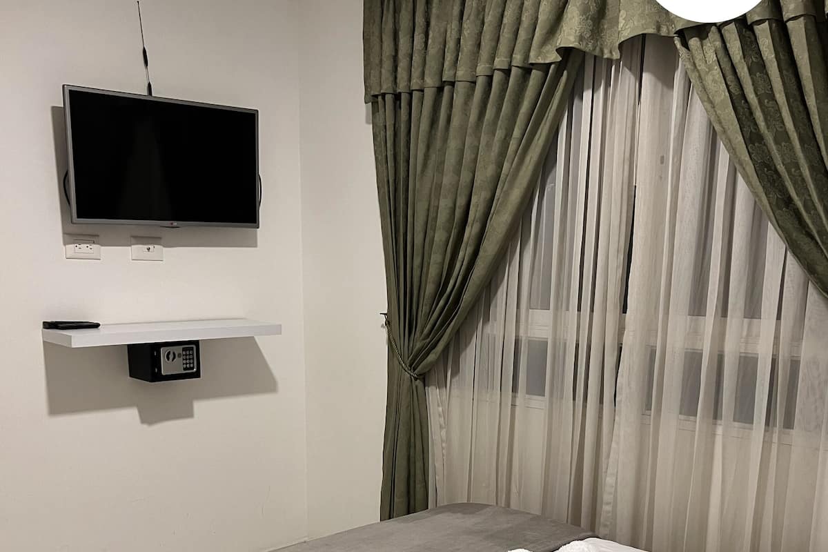 A modern hotel room with a bed, a TV mounted on the wall, and a window with sheer curtains.
