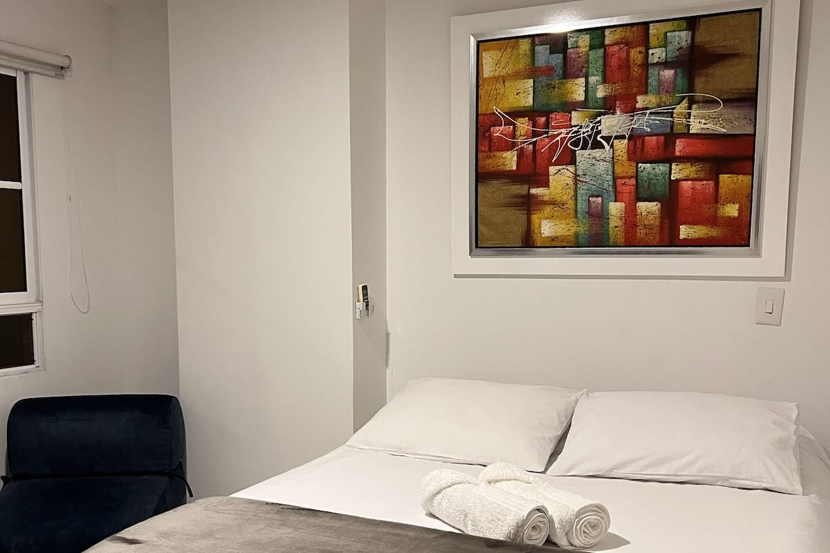 A hotel room with a bed, a blue chair, a colorful abstract painting, and a sign with 'Casa Alom' logo.