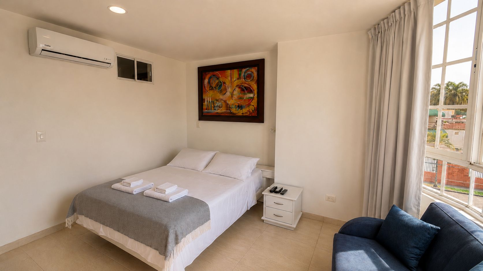 A hotel room with a bed, a bedside table, a chair, a window with curtains, an air conditioning unit, and a painting on the wall.