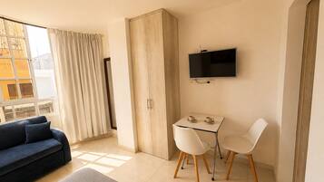 Una habitación moderna con sofá, una mesa y sillas, televisor y un amplio ventanal con cortinas.