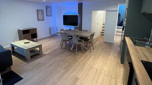 Un salon moderne comprenant un coin repas, un plancher en bois, un téléviseur à écran plat fixé au mur et un coin cuisine avec un évier et des placards.