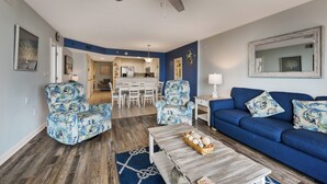 Un salon avec un canapé bleu, deux fauteuils à motifs, une table basse en bois et un ventilateur de plafond.