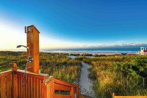 Una terraza de madera con ducha, un sendero arenoso que lleva a la playa y una torre de salvavidas al fondo.