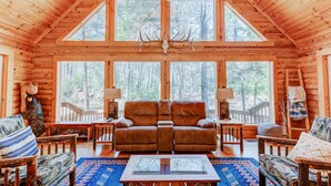 L’intérieur d’une cabane en rondins, avec une grande fenêtre, une cheminée et un coin salon confortable.