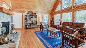 L’intérieur d’une cabane en rondins, avec une cheminée en pierre, des étagères en bois, un canapé en cuir et de grandes fenêtres.