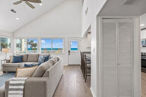 A spacious living area with a large window overlooking the ocean, a ceiling fan, and a kitchen area with a sliding door. - Eastside Wave - 5BR/3BA Gulf Trace Home - Private Pool - Sleeps 12 (Grayton)