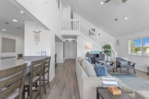 A spacious living room with a high ceiling, a staircase, and a dining area with wooden chairs and a table. - Eastside Wave - 5BR/3BA Gulf Trace Home - Private Pool - Sleeps 12 (Grayton)