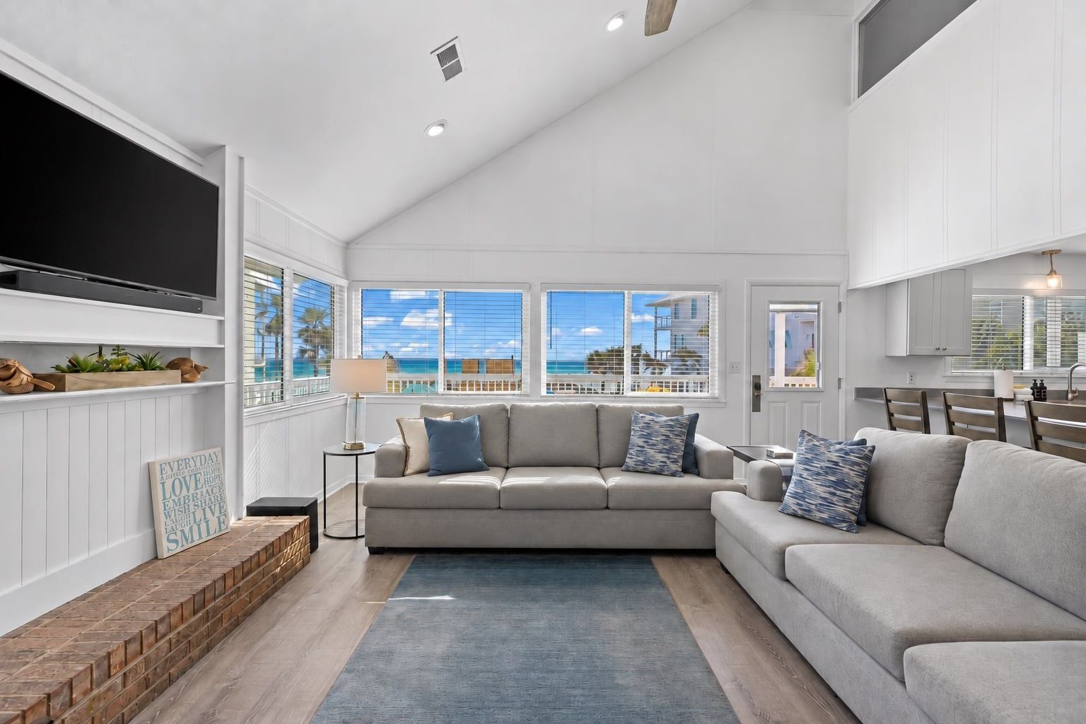 A spacious living room with a large grey sofa, a coffee table, and a TV mounted on the wall. There are large windows offering a view of the ocean.