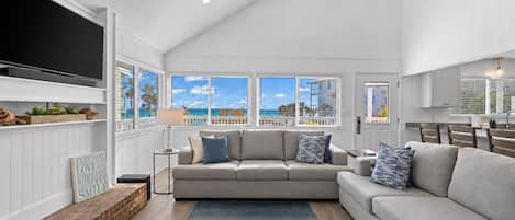 A spacious living room with a large grey sofa, a coffee table, and a TV mounted on the wall. There are large windows offering a view of the ocean.
