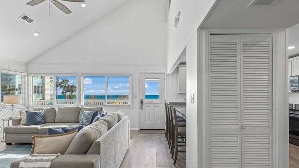 A spacious living room with a large window overlooking the ocean, a ceiling fan, and a dining area with chairs and a table.
