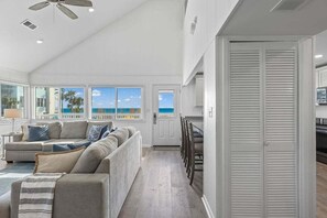 A spacious living room with a large window overlooking the ocean, a ceiling fan, and a dining area with chairs and a table. - “Eastside Wave” - Gulf Trace - Private Pool - Sleeps 12 - 100 Steps to the Gulf (Grayton)