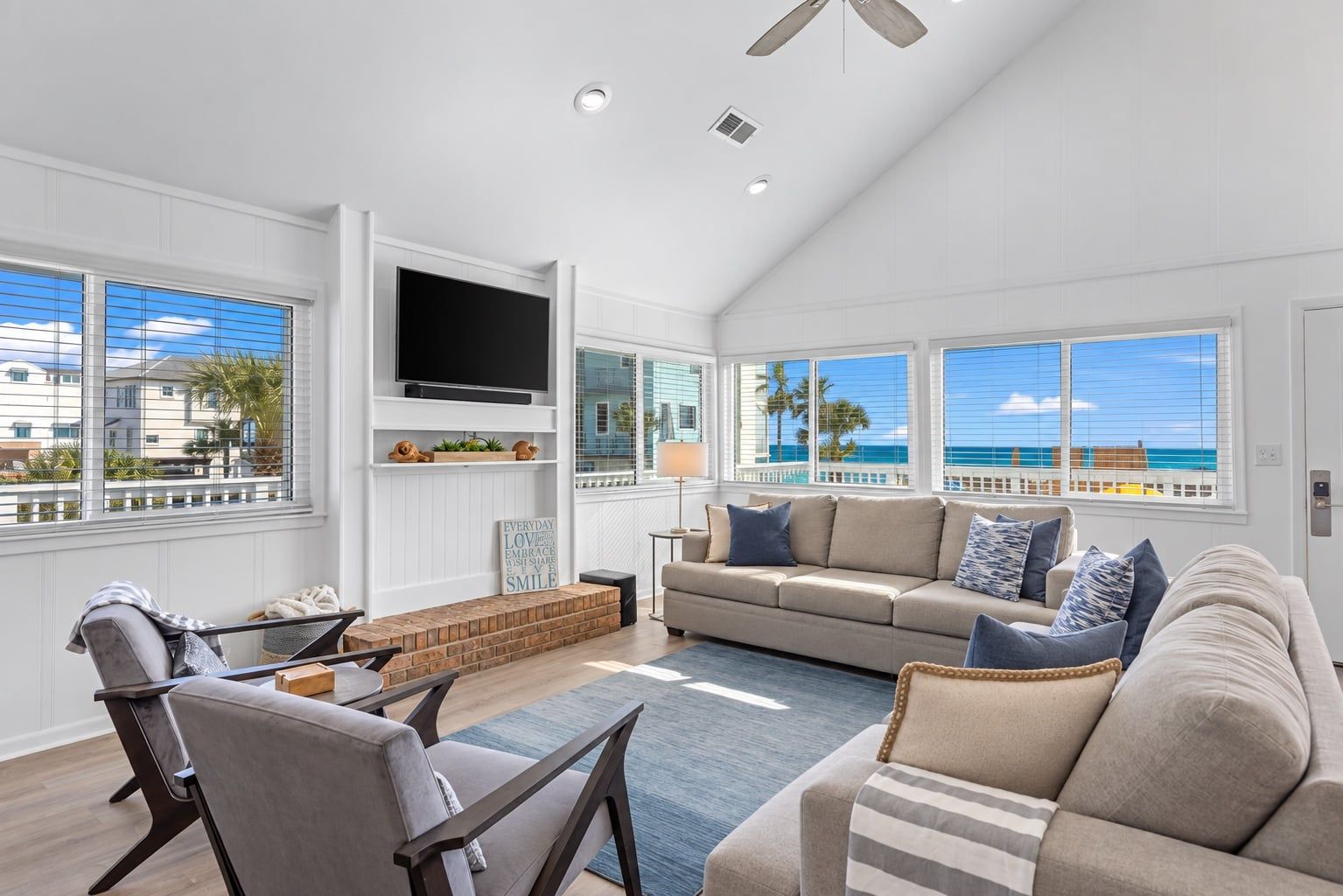A spacious living room with a large window overlooking the ocean, a flat-screen TV mounted on the wall, and a comfortable seating area with a sofa and chairs.