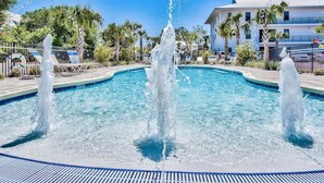 A swimming pool with multiple water jets, surrounded by palm trees and a building with balconies.