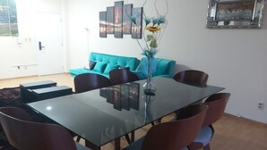 Uma área de refeições com mesa de vidro, cadeiras azuis e um sofá azul-esverdeado ao fundo.