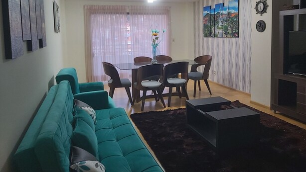 Uma sala de estar com um sofá verde-água, uma mesa de jantar com cadeiras, uma televisão e obras de arte nas paredes.