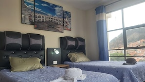Quarto de hotel com duas camas, uma mesa de cabeceira, luminária fixada na parede e uma grande janela com vista para edifícios e colinas.