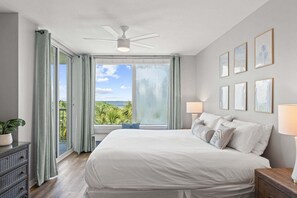 Un dormitorio con una cama grande, ventilador de techo, ventana con vista al océano y cuadros en la pared.